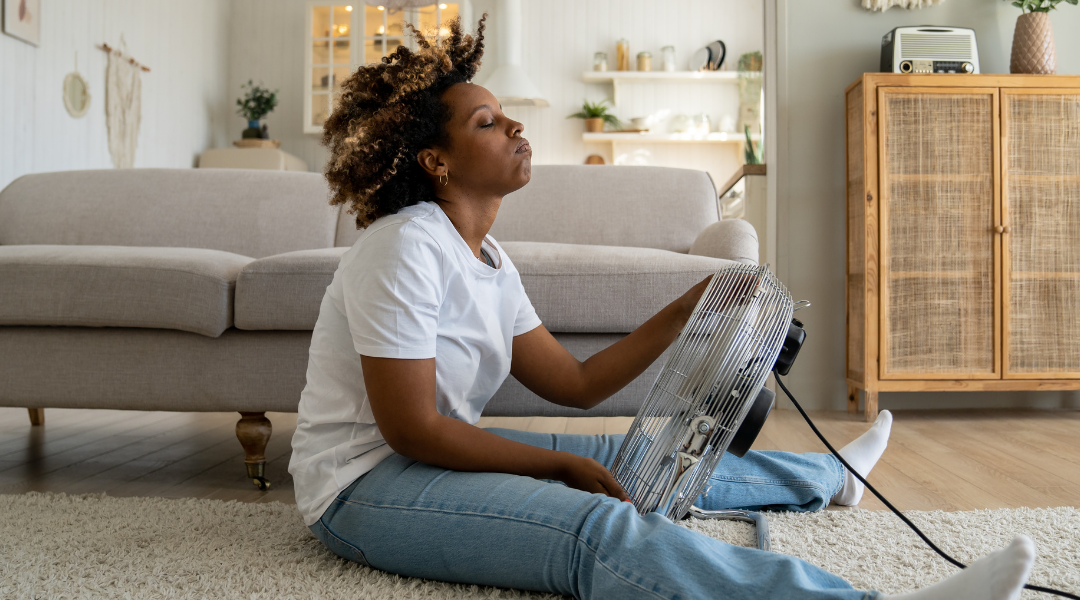 Overheated woman sitting on floor of living room trying to cool down with fan directly under her.